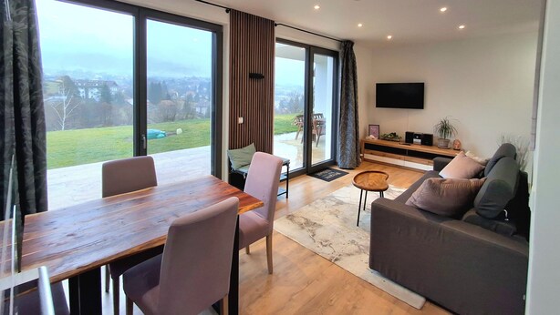 Un vaste espace de vie comprenant une grande porte-fenêtre coulissante donnant sur un balcon, une table à manger en bois avec des chaises, un canapé gris et un téléviseur à écran plat fixé au mur.