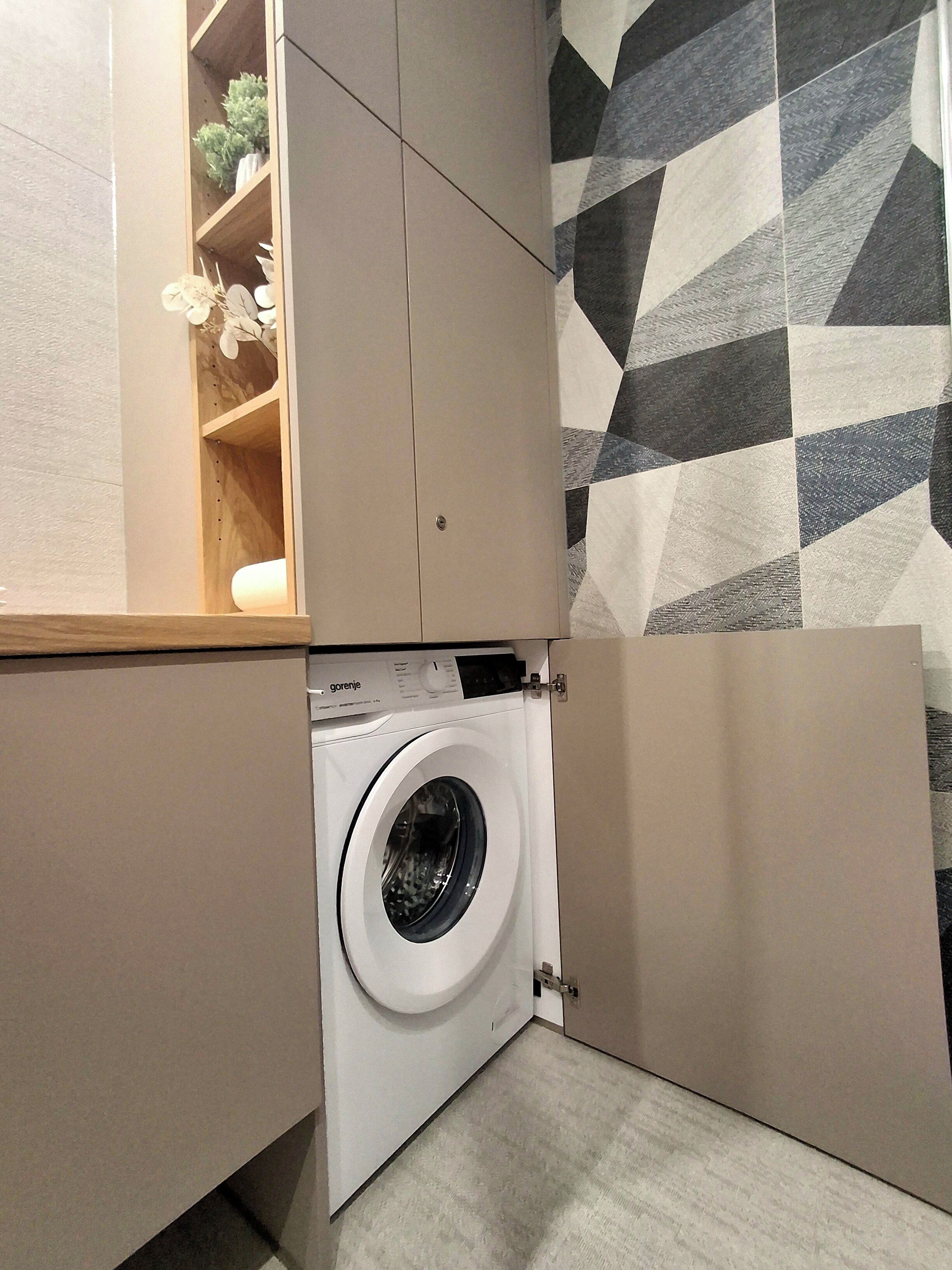 A compact laundry room with a washing machine, wooden shelves with plants, and a geometric-patterned wall.