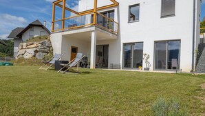 Une maison moderne à deux étages, dotée d’un balcon, d’un mur en pierre et d’une cour verdoyante avec des chaises longues.