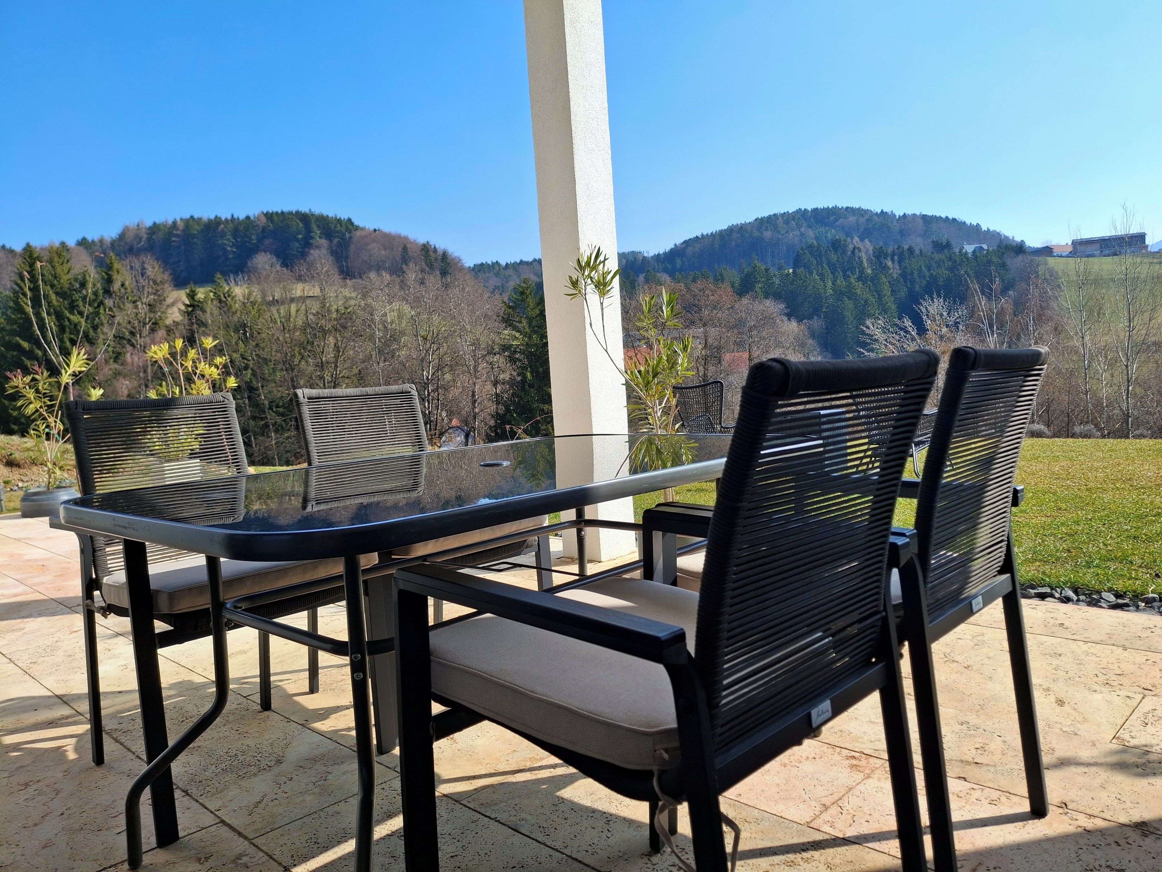 A patio with a glass table and chairs, a white pillar, and a view of a forested hillside.