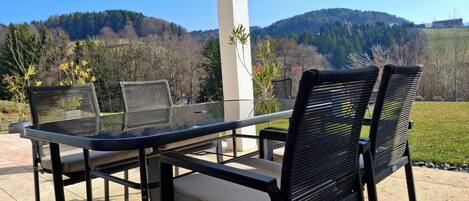 A patio with a glass table and chairs, a white pillar, and a view of a forested hillside.