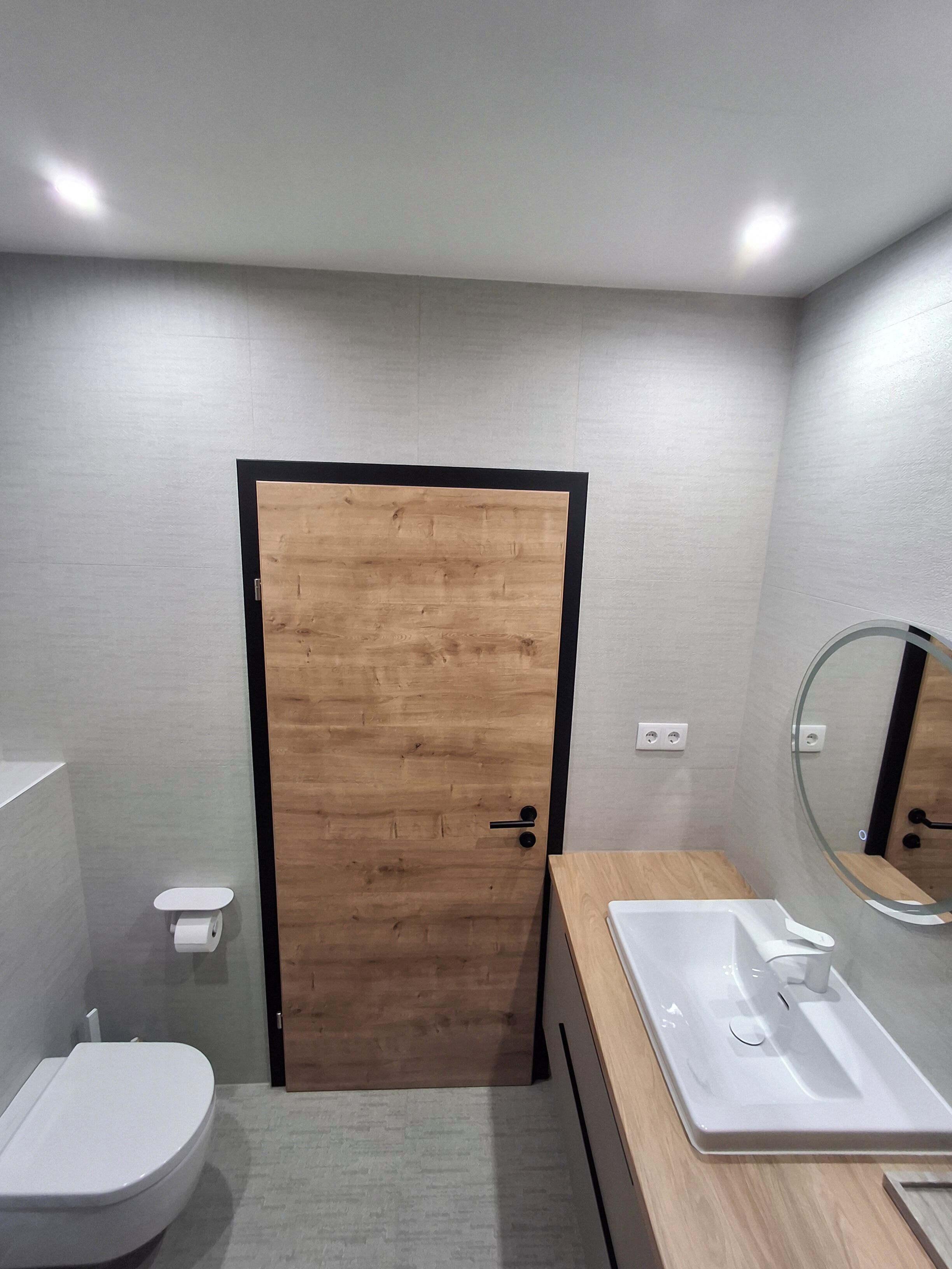 Une salle de bain moderne avec une porte en bois, des toilettes blanches, un lavabo avec un miroir et une petite étagère.