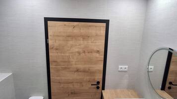 Une salle de bain moderne avec une porte en bois, des toilettes blanches, un lavabo avec un miroir et une petite étagère.