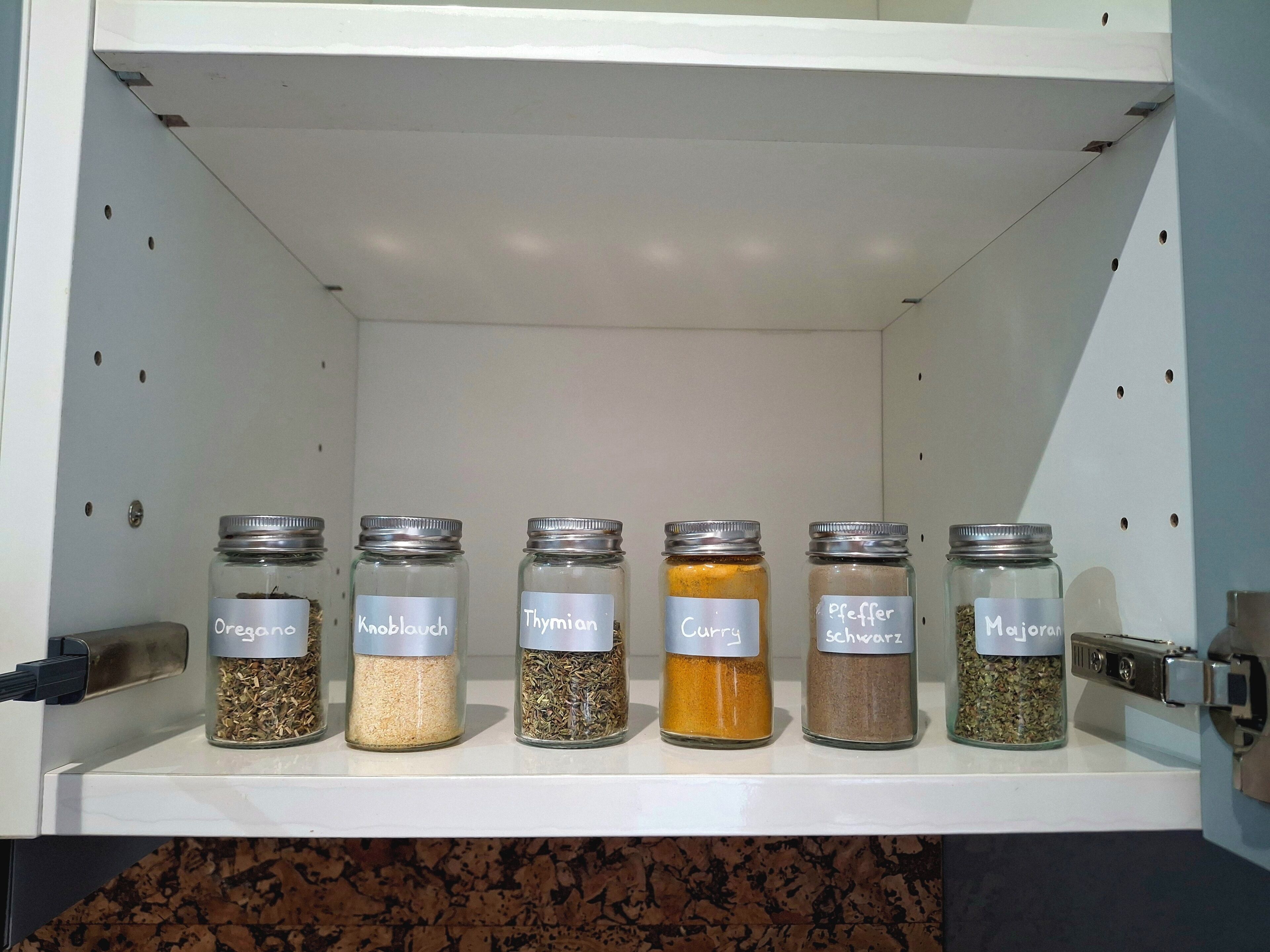 Une étagère sur laquelle sont disposés six bocaux en verre contenant différentes épices, chacun étant étiqueté avec son nom.