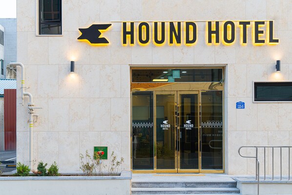 Esterno dell'Hotel Hound con un cartello ben visibile, porte in vetro e un piccolo cartello verde con simboli.