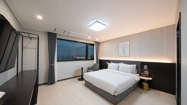 Quarto de hotel moderno com uma cama grande, uma mesa, uma cadeira e uma pequena mesa com abajur.