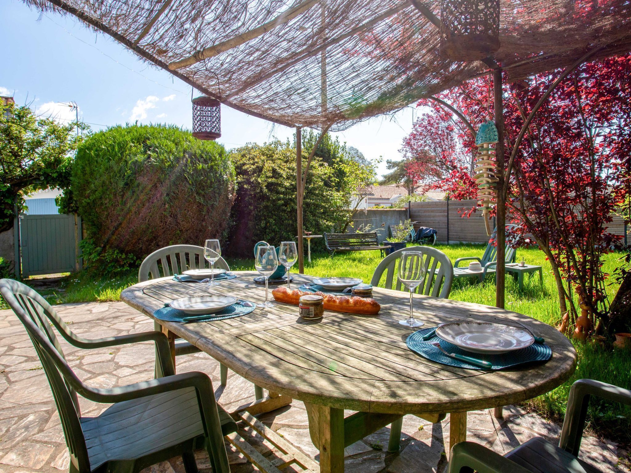 Een houten eettafel buiten, gedekt met stoelen, met een geweven luifel erboven en een tuin op de achtergrond.