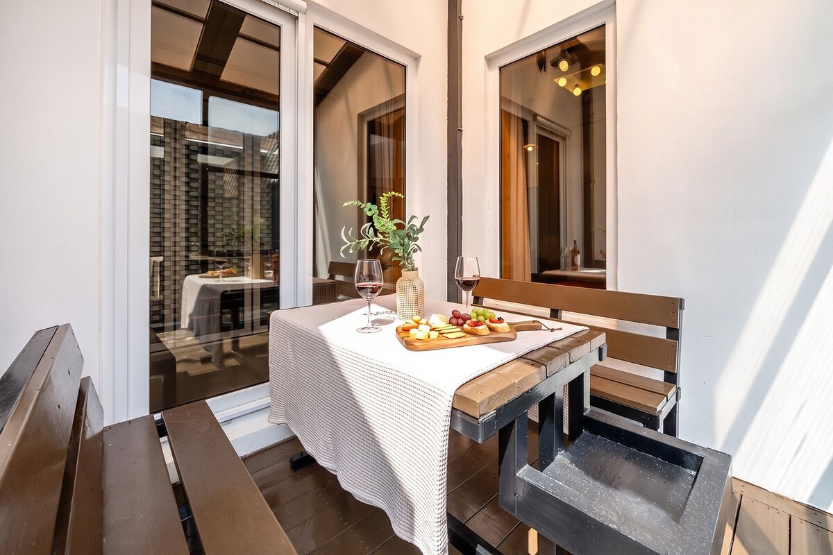 An outdoor dining area with a table set for two, a wooden bench, and a view of a restaurant through the glass door.