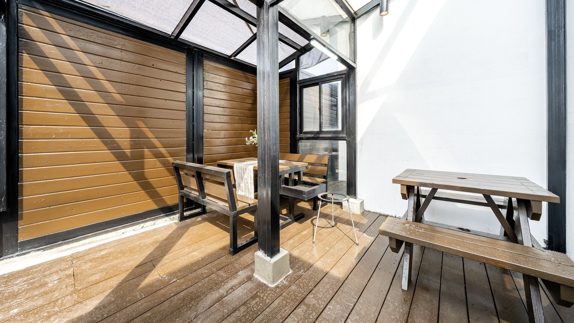 Um pátio externo com piso de madeira, uma mesa, bancos e um compartimento envidraçado.