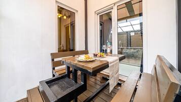 An outdoor wooden deck with a table and benches, a glass door leading to a patio, and a view of a brick wall.