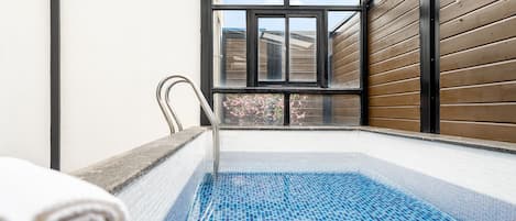 A small indoor pool with a tiled floor, a metal chair, and a view of a wooden building through a window.
