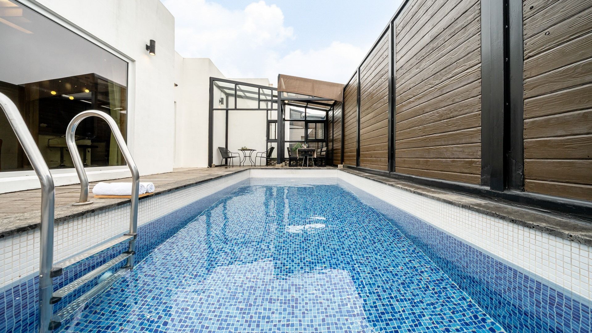 A swimming pool with a tiled floor, a metal ladder, and a seating area with tables and chairs.