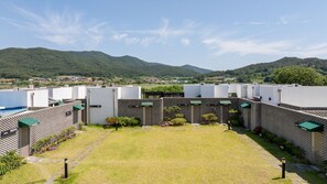 A courtyard with a central lawn, surrounded by buildings with external air conditioning units, and a mountainous backdrop. - Buan Cordelia Pool Villa Pension (Buan)