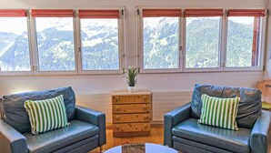 A cozy living room with two leather sofas, a wooden side table, and a view of snow-covered mountains through large windows.