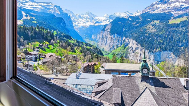 A view from a window overlooking a mountainous landscape with snow-capped peaks, a village with traditional buildings, and a clock tower.