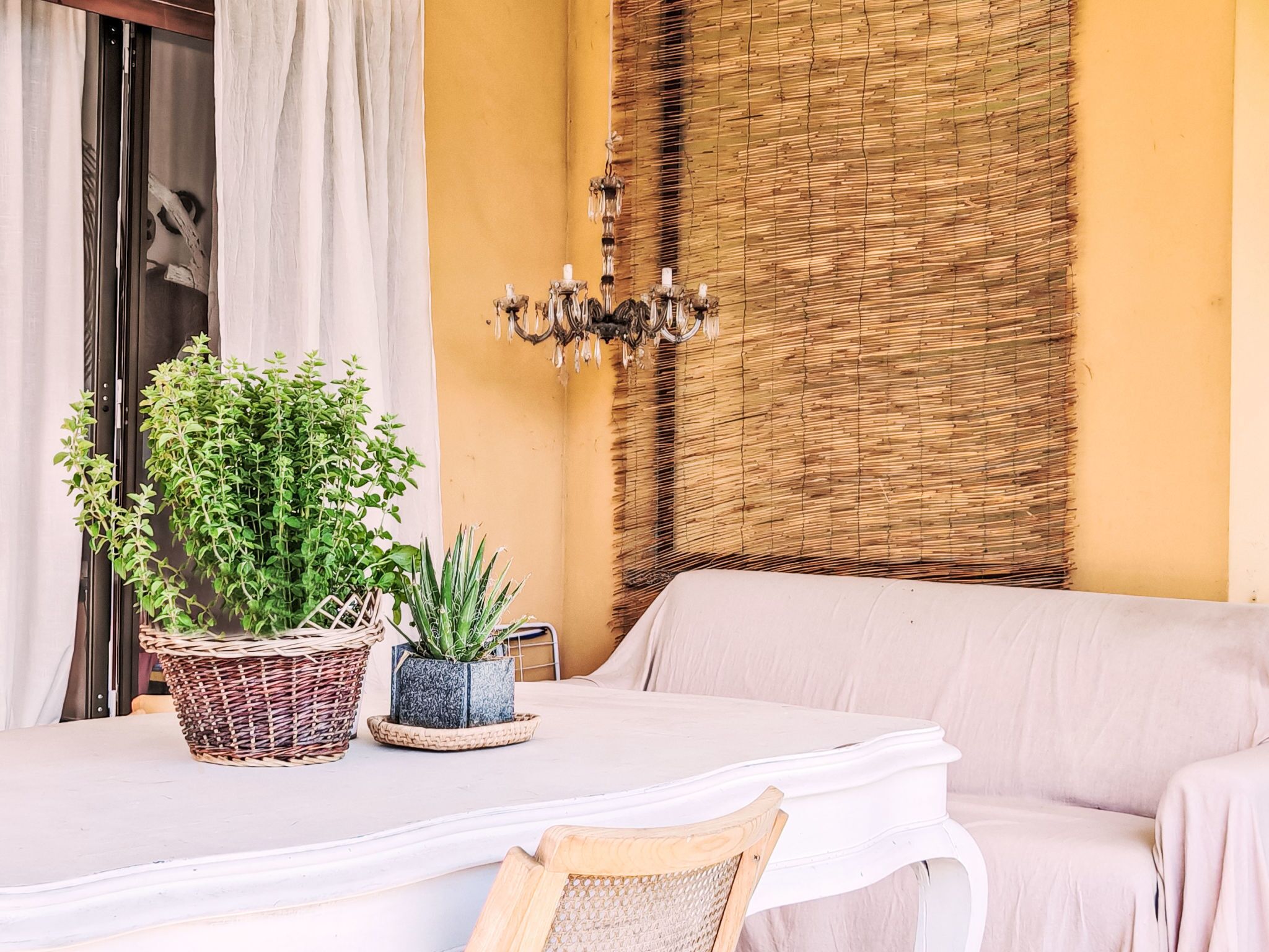 Ein Zimmer mit weißem Tisch, Korbwannen mit Pflanzen, einer Wandverkleidung aus Weide und einem weißen Sofa, sowie einer Kristallleuchter.