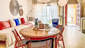 Ein gemütliches Wohnzimmer mit einem runden Holztisch, einer Couch mit bunten Kissen und einem Essbereich mit Korbstühlen.