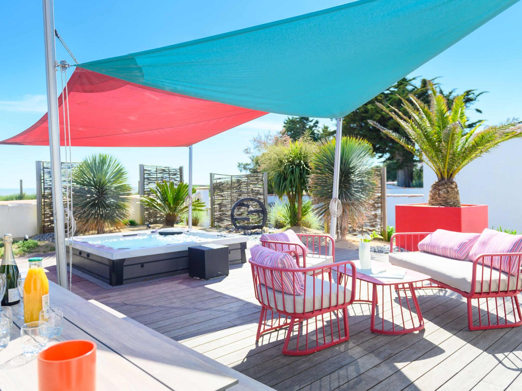 A rooftop terrace with a hot tub, red and blue umbrellas, and red wicker chairs.