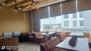 A modern restaurant with high ceilings, a patterned ceiling, and large windows offering a view of buildings outside. - Hotel Taj Royal (Agra)