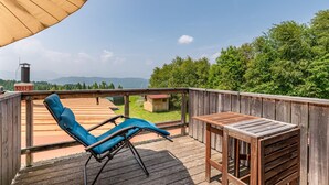 Une terrasse en bois avec un fauteuil de jardin bleu et une petite table, d’où l’on peut admirer une zone boisée avec des montagnes à l’horizon.
