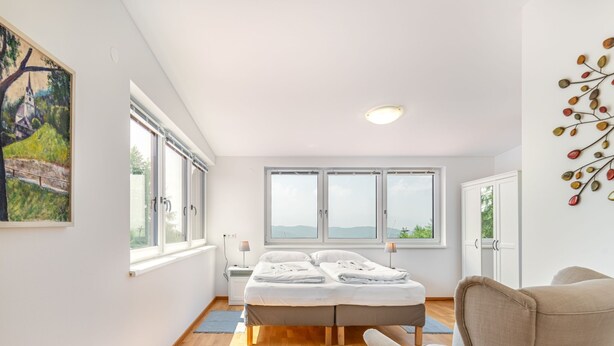 Une chambre moderne avec un grand lit, un fauteuil et une vue sur l’extérieur grâce à de grandes fenêtres.