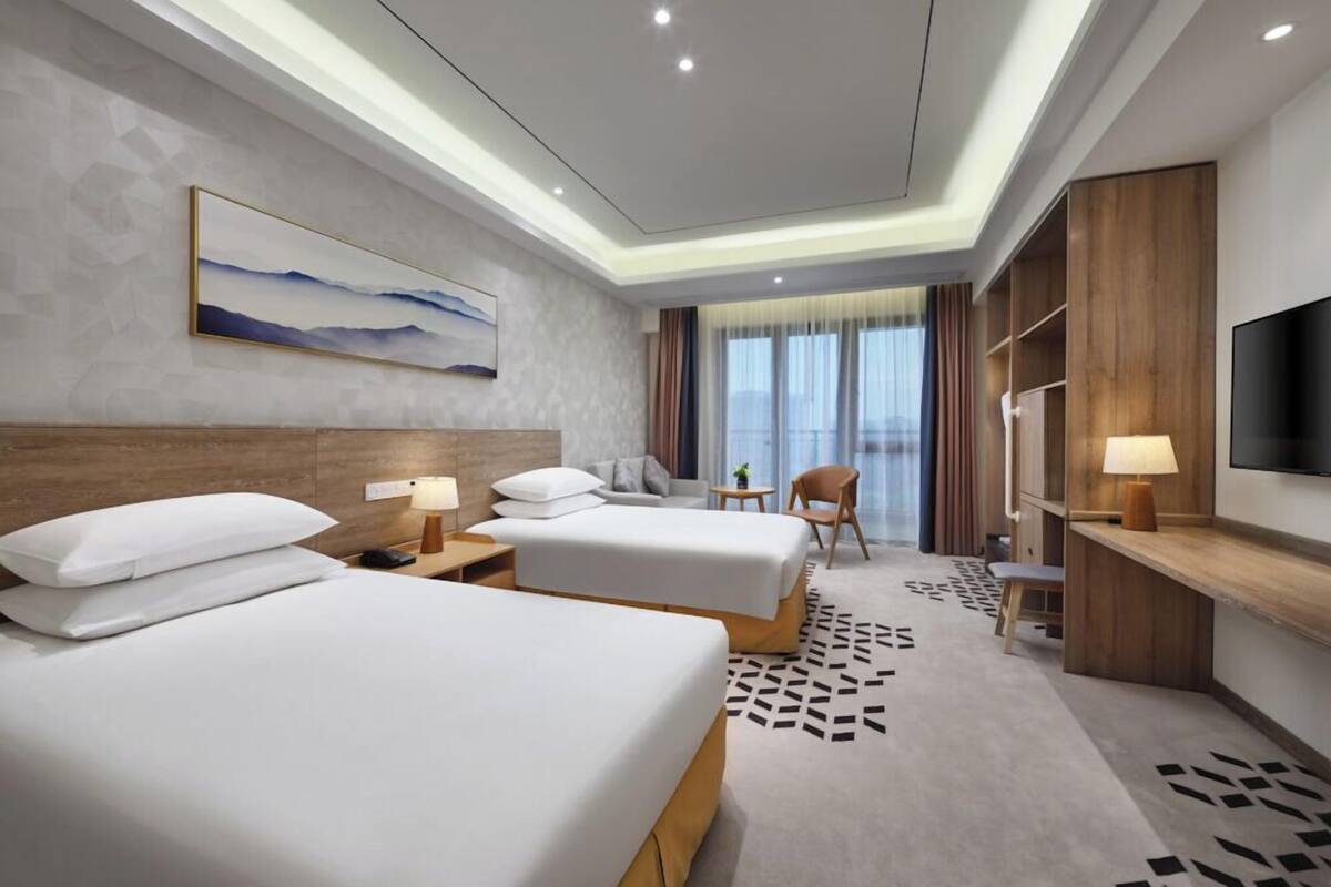 Ein Hotelzimmer mit zwei Betten, einem Schreibtisch, einem Stuhl, einem Fernseher und einem großen Fenster mit Vorhängen.