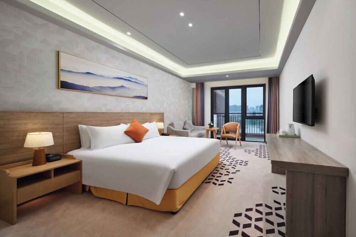 Ein modernes Hotelzimmer mit einem großen Bett, einem Schreibtisch, einem Sessel, einem Fernseher und einem Balkon mit Blick auf die Stadt.
