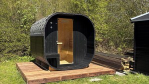 A small, black, cylindrical wooden structure with a door, situated on a wooden platform in a grassy area with trees in the background. - Lovely cottage, close to beach and forest (Nakskov)