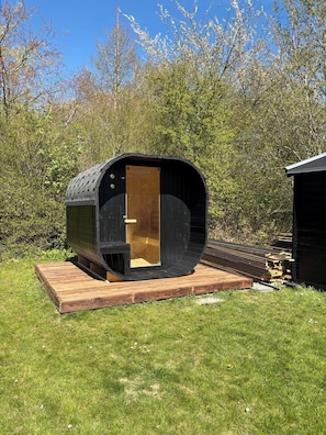 A small, black, cylindrical wooden structure with a door, situated on a wooden platform in a grassy area with trees in the background.