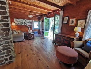 A cozy cabin interior with wooden walls, a stone fireplace, a sofa, a coffee table, and a large window offering a view of the outdoors.