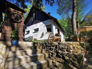 Una cabaña rústica con una escalera de piedra que lleva a una terraza de madera, rodeada de árboles y un cielo azul despejado.