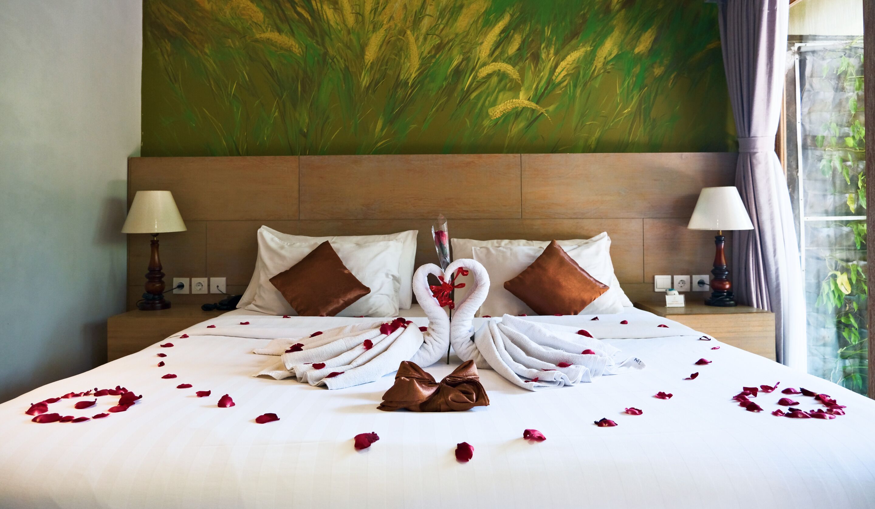 Une chambre d’hôtel avec un grand lit, du linge de lit blanc, des pétales de rose disposés en forme de cœur et un tableau au mur.