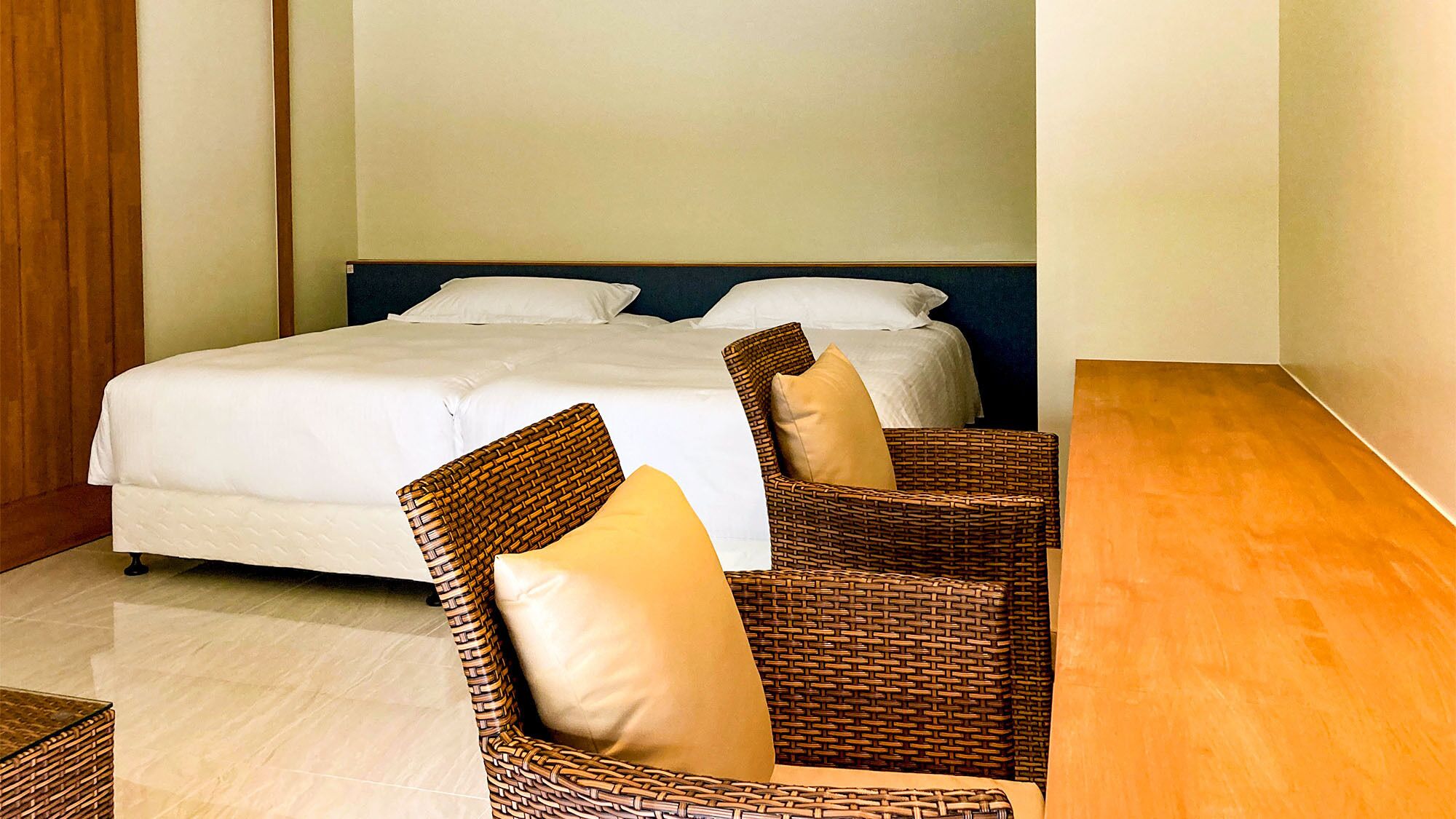 A hotel room with a bed, two wicker chairs, a wooden table, and a plain wall.