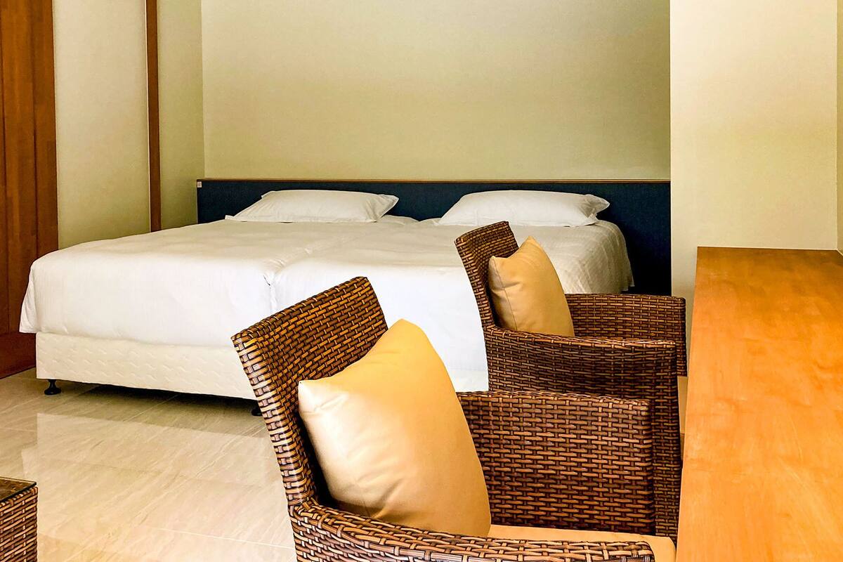 A hotel room with a bed, two wicker chairs, a wooden table, and a plain wall.