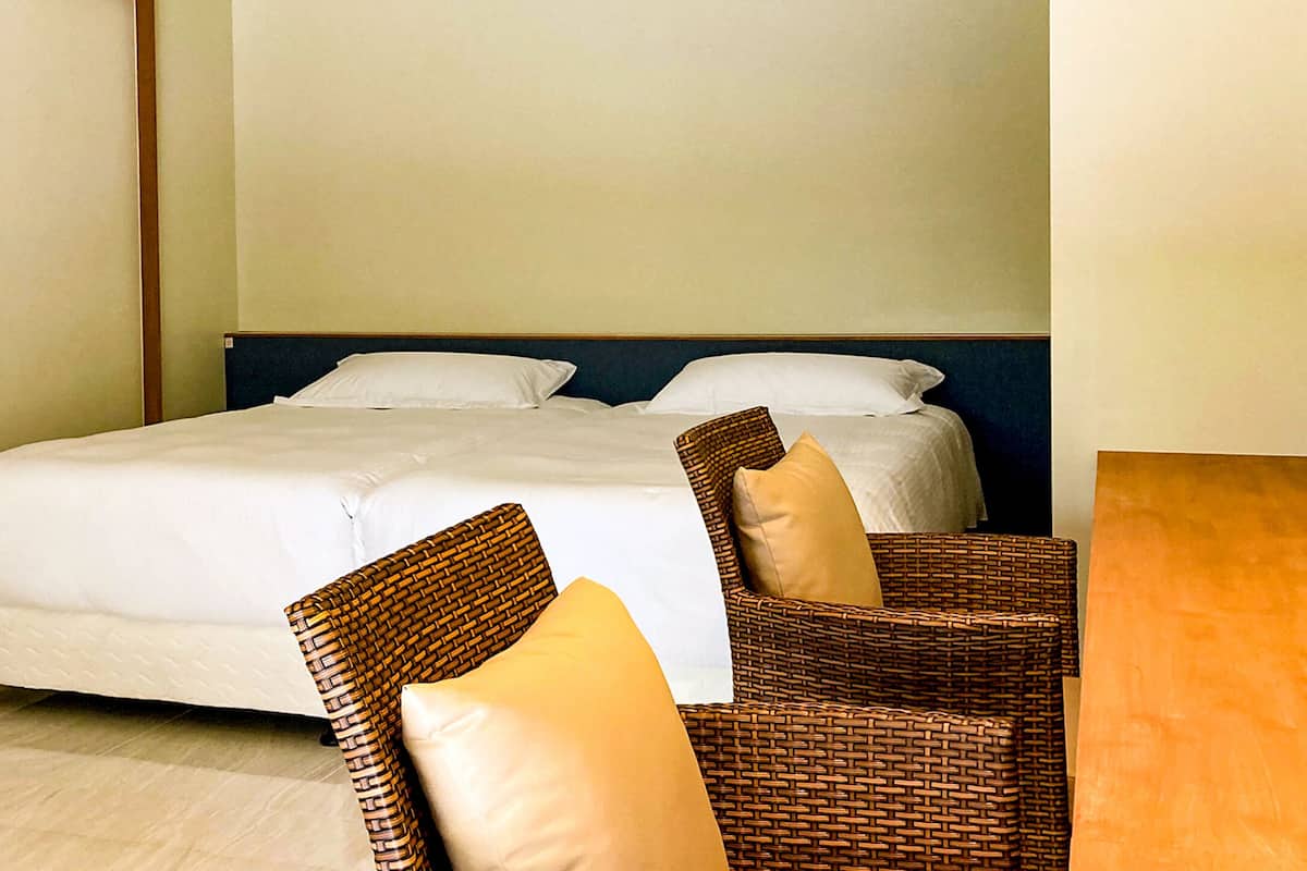 A hotel room with a bed, two pillows, a wicker chair, and a wooden table.