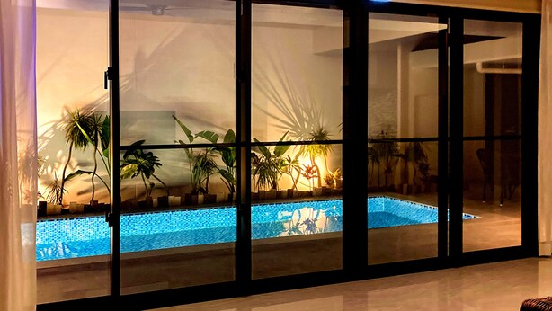 A modern indoor pool area with clear glass walls, surrounded by potted plants and a tiled floor. - villa LIFE INDAH sumuide (Nago)