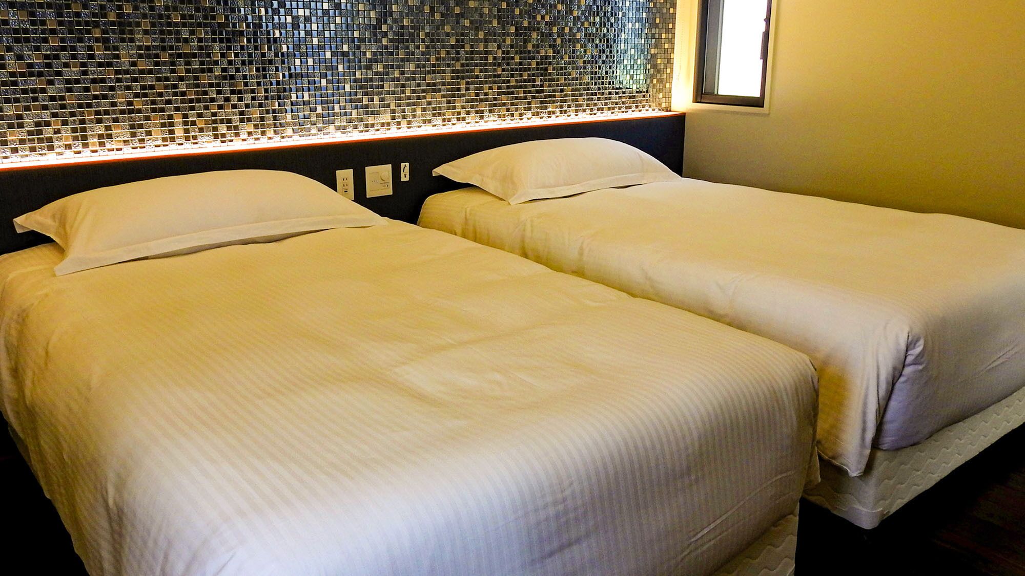 Two single beds with white linens in a room with a mosaic tile wall and a window.