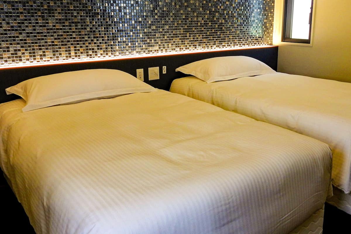 Two single beds with white linens in a room with a mosaic tile wall and a window.