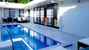 A modern outdoor pool area with a clear blue pool, a lounge with black and white chairs, and a glass-enclosed patio. - villa LIFE INDAH sumuide (Nago)
