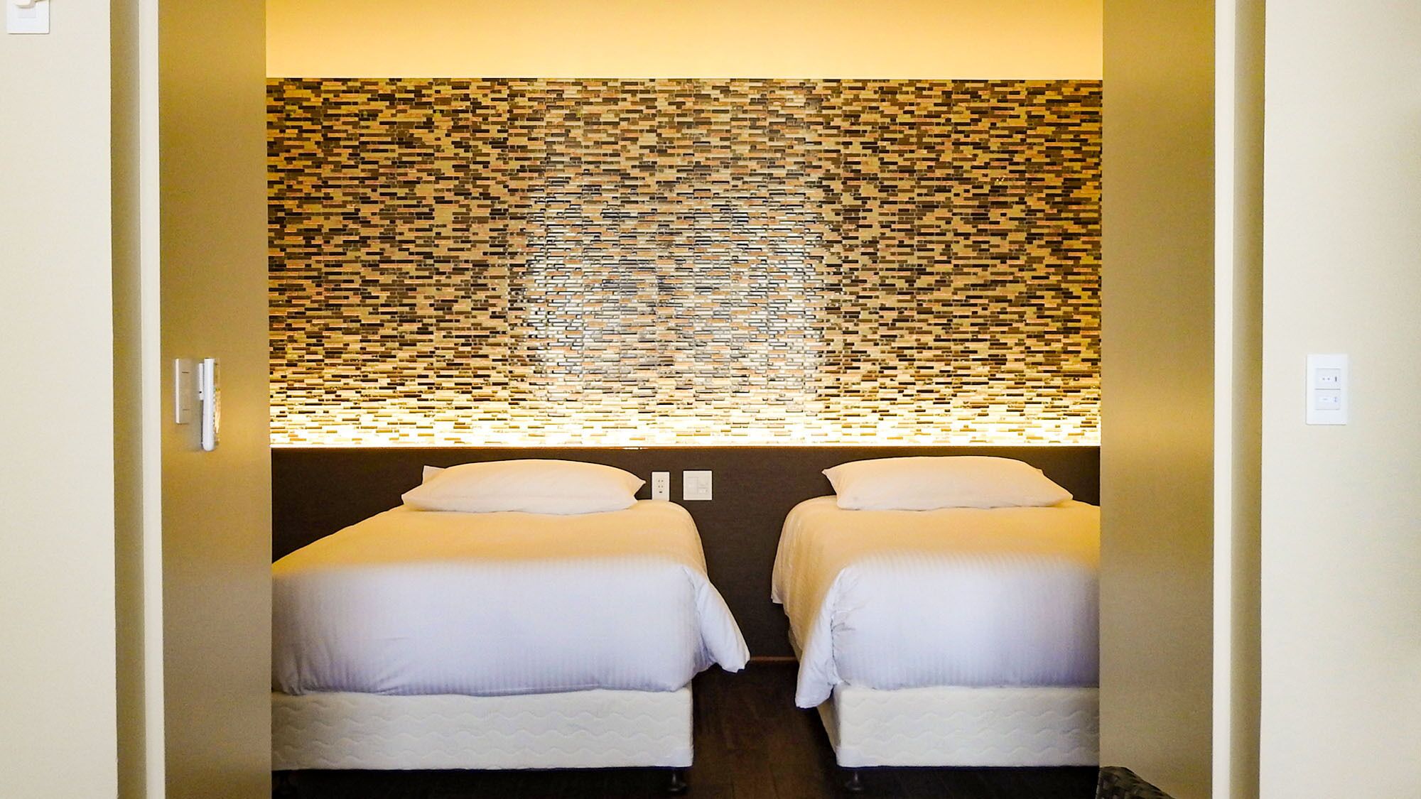 A room with two beds, a wall with a mosaic pattern, and a light fixture above the beds.