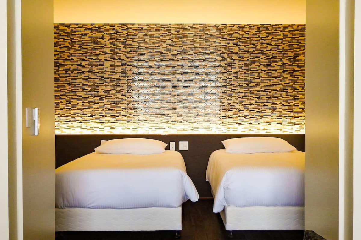 A room with two beds, a wall with a mosaic pattern, and a light fixture above the beds.
