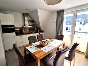 A modern kitchen with white cabinets, a dining table with four chairs, and a view of a residential area through a large sliding glass door. - Beachfront apartment with balcony, pool, and sauna - Rex Rugia - Rex Rugia (Göhren)