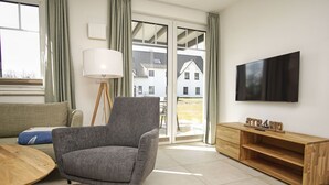 A modern living room with a grey armchair, a wooden coffee table, a TV on the wall, and a sliding glass door leading to a balcony. - Beachfront apartment with terrace, use of swimming pool and sauna - Rex Rugia H-1010 - (Göhren)