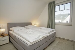A double bed with white linens, a bedside table with a lamp, and a window with curtains. - Beachfront vacation rental with a balcony, access to the pool and sauna - Rex Rugia H-1010 - (Göhren)