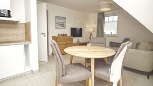 A modern living room with a round wooden dining table, grey upholstered chairs, a grey sofa, a wooden cabinet, a television, and a painting on the wall. - Beachfront vacation rental with a balcony, access to the pool and sauna - Rex Rugia H-1010 - (Göhren)