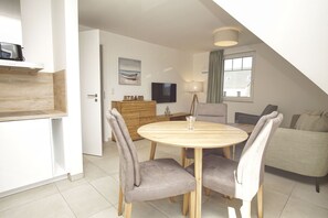 A dining area with a round wooden table and four grey upholstered chairs, a wooden cabinet, a television, and a painting on the wall. - Beachfront vacation rental with a balcony, access to the pool and sauna - Rex Rugia H-1010 - (Göhren)