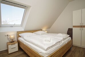 A neatly made bed in a room with a sloped ceiling, a single bedside table with a lamp, and a wardrobe.