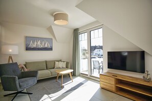 A modern living room with a sofa, armchair, TV, and a painting of sailboats. - Beachfront vacation rental with a balcony, access to the pool and sauna - Rex Rugia - Rex Rugia (Göhren)