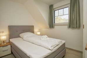 Ein ordentlich bezogenes Bett in einem Zimmer mit geneigter Decke, einem Fenster mit Vorhängen und Nachttischlampen.