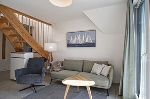 A living room with a wooden staircase, a sofa, a chair, a coffee table, and a painting of sailboats. - Beachfront vacation rental with a balcony, access to the pool and sauna - Rex Rugia - Rex Rugia (Göhren)